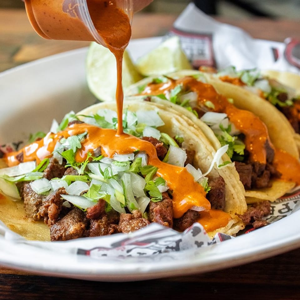 Sauce being poured over three El Paisa Cocina Mexicana tacos with meat, lettuce, and onions.