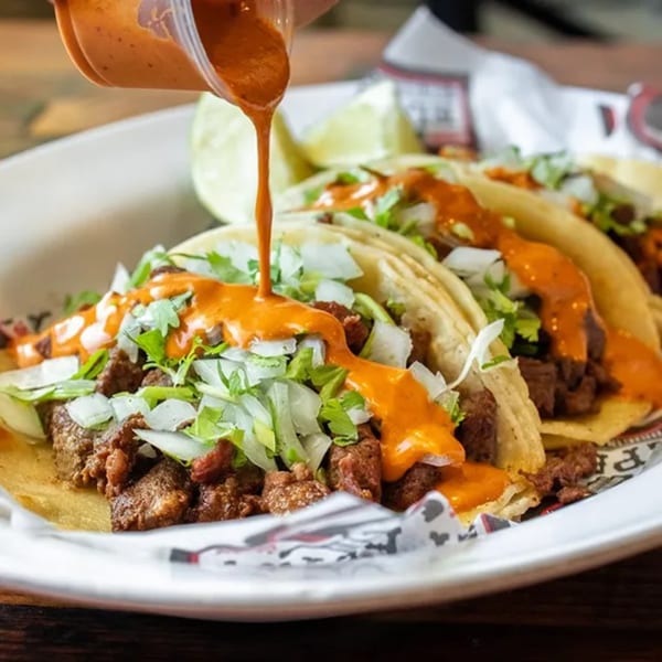 Savory El Paisa Cocina Mexicana tacos with meat, onions, cilantro, and red sauce being poured.