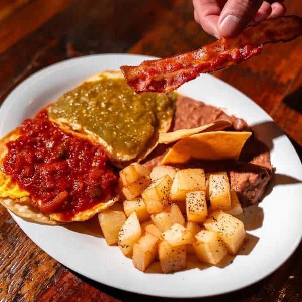 A hand holds a crispy bacon strip over a Mexican breakfast plate with eggs, salsa, beans, and potatoes.